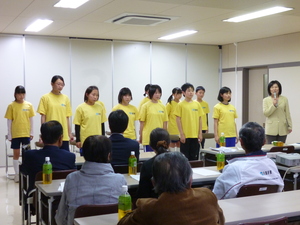 マイクを持って話している女性の横で、黄色いシャツを着た学生が横に並んでいる写真