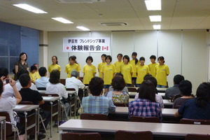 体験報告会で、黄色いシャツを着た学生たちが、着席している男女の前に並んでいる写真