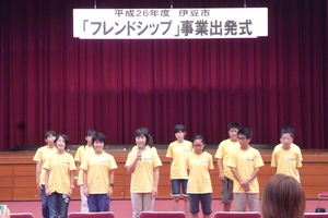 お揃いの黄色いTシャツを着た生徒たちが2列に並んでステージ前に立ち、前列中央にいる女の子が発表をしている様子の写真