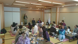 料理が並べられた席についた参加者の方々が、前方に立つ外国人の女性の話を聞いている様子の写真