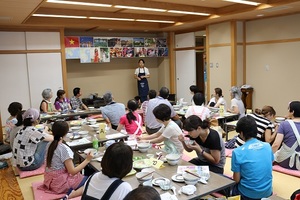 座敷で4か所に分かれて座り、食事をとりながら前に立つ講師の話を聞いている参加者の方々の写真