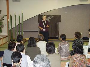着物や竹飾りが飾られた和室で語り手の女性が立って話をしているのを座って聞いている参加者の方々の写真