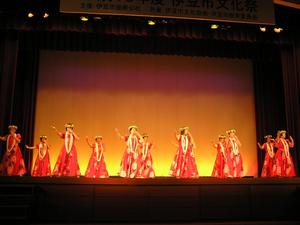 首や頭に花輪を付け、赤いドレスを着た10名ほどの女性がステージ上でフラダンスを披露している様子の写真
